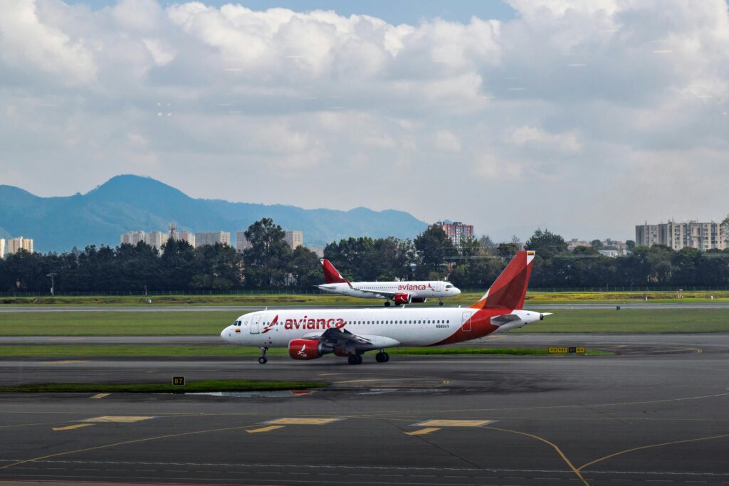 Two Avianca Airlines planes taxi on a runway of an unidentified airport with mountains and partly cloudy skies in the background.