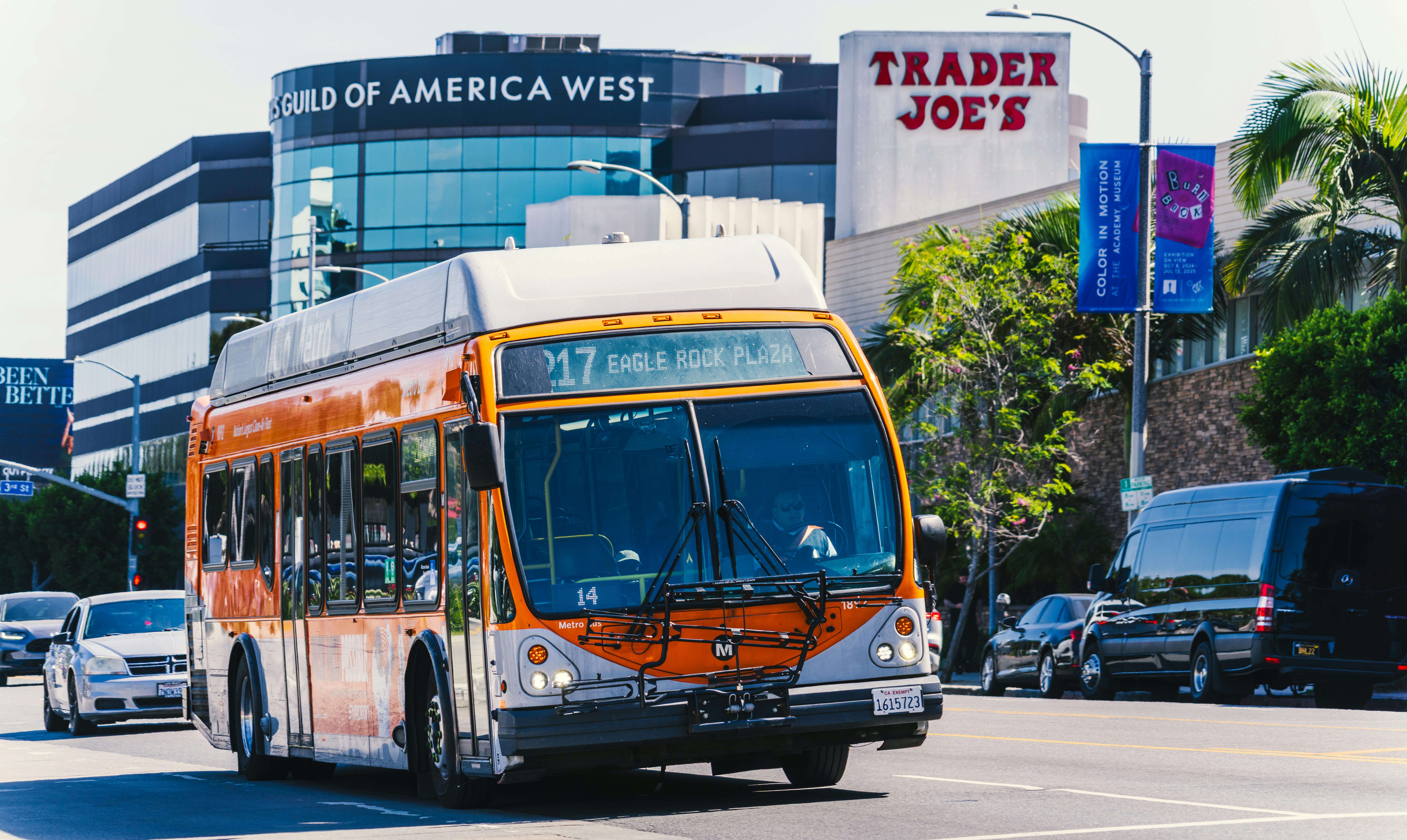 An orange LA Metro bus travels on Los Angeles surface streets in front of a Trader Joe's grocery store as gas prices soar